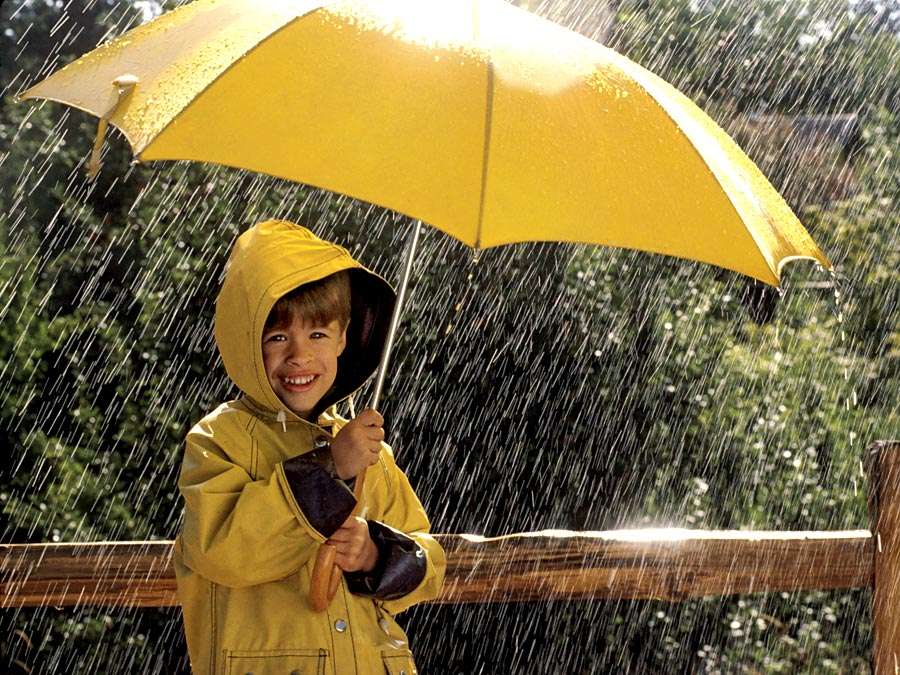 Boy in yellow raincoat with hood under a yellow umbrella on a rainy day.